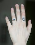 A close-up of a person's hand showing two rings: a silver band with a round light blue gemstone on the ring finger, and a small silver geometric ring on the middle finger. The background is a dark, blurred gray.