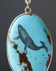 An oval pendant necklace featuring a light blue turquoise stone with natural brown matrix and a detailed black whale illustration, set in a gold-tone bezel with a matching chain.