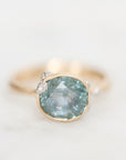 A gold ring featuring a large oval light blue-gray gemstone with small clear accent stones on the shoulders, set against a soft white background.