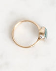 A gold ring with a small oval light blue gemstone and tiny clear accent stones, photographed on a light marble-like surface.