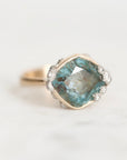 A cushion-cut labradorite ring with a blue-green iridescent center stone, surrounded by a silver halo with small clear accent stones, set on a gold band, photographed on a light marble surface.