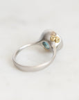 A silver ring with a textured, organic-shaped top featuring two small gold flower-like details and a tiny light blue stone, shown on a light marble surface.