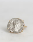 A gold ring featuring an antique silver coin with an embossed profile portrait, set against a plain light gray background.
