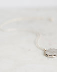 A silver necklace with a round pendant featuring a textured, frosted white stone, resting on a light marble surface.