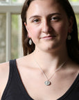 A woman with long brown hair wearing a silver pendant necklace and matching silver earrings, photographed indoors with a softly blurred window background.
