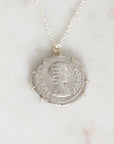 A silver pendant necklace featuring an ancient coin pendant with a profile portrait, suspended from a delicate silver chain on a light marble background.