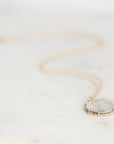 A delicate gold chain necklace with a round, textured silver pendant resting on a light marble surface.