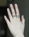 A close-up of a person's hand wearing a delicate gold ring with a small oval yellow gemstone on the ring finger, with a small geometric tattoo on the middle finger, against a dark blurred background.