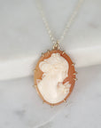 An oval cameo pendant necklace featuring a carved profile of a woman on a peach and cream shell, set in a gold-toned bezel with small spikes, hanging from a silver-toned chain on a white marble surface.