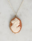 A vintage-style oval pendant necklace featuring a cream-colored carved classical profile on a peach-orange background, set in a gold-toned bezel with small spike details, hanging from a silver-toned chain on a light marble surface.