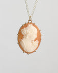 A vintage-style oval pendant necklace featuring a cream-colored carved classical profile on a peach-orange background, set in gold-toned metal with a delicate chain against a light gray background.