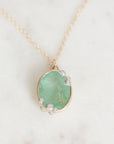 An oval pendant necklace featuring a light green stone in a gold bezel setting with small silver accents, resting on a white marble surface.