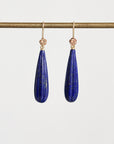 A pair of gold drop earrings with teardrop-shaped dark blue lapis lazuli stones, hanging from a gold rod against a light gray background.