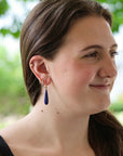 A woman wearing a dark blue teardrop-shaped drop earring with gold hoop and stud earrings, shown in a close-up profile against a blurred green outdoor background.