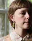 A close-up portrait of a woman wearing a pair of dangle earrings made of two stacked, rounded olive-green beads on a gold-toned hook, set against a soft-focus background of a window.