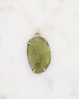 An oval green stone pendant necklace with a gold-toned bezel setting and delicate chain, displayed on a light marble surface.