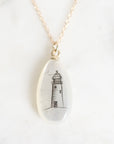 A gold chain necklace with an oval clear resin pendant featuring a black ink sketch of a lighthouse, displayed on a light marble surface.