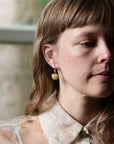 A close-up portrait of a woman wearing a small, round, light green bead earring with a gold-toned top hook, paired with a cream-colored patterned blouse.