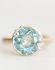 A round-cut light blue gemstone ring in a gold-toned setting, accented with three small clear stones, photographed on a soft light gray background.
