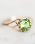 A gold ring featuring a round-cut green peridot gemstone in a prong setting, resting on a light marble surface.