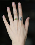A close-up of a person's hand wearing a gold ring with a green gemstone, set against a dark blurred background.