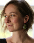 A woman with short brown hair is shown in profile, wearing a dangling green teardrop-shaped earring. The photo is taken indoors near a bright window, with soft natural light and a blurred light background.