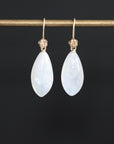 A pair of teardrop-shaped white moonstone drop earrings with gold hooks, hanging from a gold display bar against a solid black background.