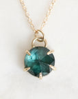 A gold necklace with a faceted round dark teal gemstone pendant in a prong setting, shown on a light off-white background.