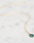 A delicate gold chain necklace with a small, faceted teal-green gemstone pendant in a gold bezel setting, resting on a light marble surface.
