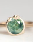 A close-up shot of a gold ring featuring a round, faceted green gemstone with natural inclusions, set in a textured gold bezel with small accent stones on top, on a light gray background.