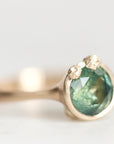 A close-up shot of a gold ring featuring a round faceted green gemstone in a prong setting, with two small accent stones on the shoulders, set against a soft, out-of-focus light gray background.