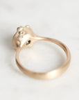 A close-up of a gold ring with a sculptural, organic-shaped top featuring a tiny green stone, resting on a light marble surface.