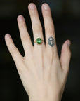 A close-up of a hand wearing a gold ring with a green oval gemstone, set against a dark blurred background.