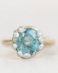 A round-cut light blue gemstone ring set in a yellow gold halo with small clear accent stones, photographed on a clean white background.