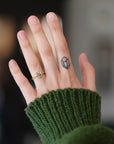 Hand with tattoo wearing gold ring with green center stone. She is wearing a green sweater against a blurred background.