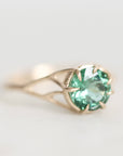 A gold ring featuring a round-cut light green gemstone in a prong setting, photographed on a clean, light gray background.