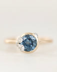 A yellow gold ring featuring a round-cut blue gemstone in a bezel setting, accented by small clear barnacle details on the shoulders, set against a soft white background.