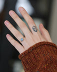 A close-up of a person's hand wearing a delicate gold ring with a round blue gemstone, set against a blurred background with a rust-colored knit sweater sleeve visible.