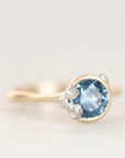 A close-up shot of a gold ring featuring a round-cut light blue gemstone accented by three small barnacle details, set against a soft, light off-white background.