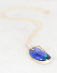A teardrop-shaped pendant necklace featuring a vibrant blue iridescent stone in a gold bezel setting, suspended from a delicate gold chain on a light marble background.