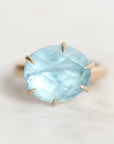 An oval-cut light blue gemstone ring in a gold prong setting, photographed on a clean, light off-white background.