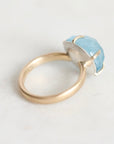 A minimalist ring featuring a polished gold band and a bezel-set light blue stone, photographed on a clean, light off-white surface.