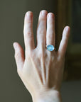 A close-up of a person's hand wearing a silver ring with a round light blue gemstone, set against a softly blurred indoor background.