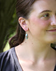 A woman wearing a dangling teardrop earring with a textured, glittery light blue-green stone in a gold-toned setting, photographed outdoors against a blurred green background.