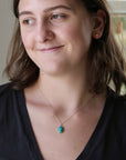 A woman with brown hair wearing a delicate gold chain necklace with a small oval turquoise pendant, paired with a black V-neck top.