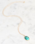 A delicate gold chain necklace with an oval turquoise pendant featuring black inlay details, resting on a white marble surface.