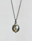 A dark gunmetal chain necklace with a pendant featuring a large dark gray pearl and a small gold-colored bird charm, set against a plain light gray background.