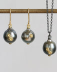 A jewelry set featuring a necklace and matching drop earrings with dark gray baroque pearls accented by gold leaf, displayed on a gold-colored rod against a light gray background.