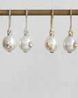 Four pearl drop earrings hanging from a gold-toned rod against a light gray background. Two earrings have white freshwater pearls with small silver floral accents, and two have white pearls with small gold floral accents.