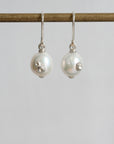 A pair of silver drop earrings with large white pearl beads and small silver floral embellishments, hanging from a gold-toned rod against a light gray background.